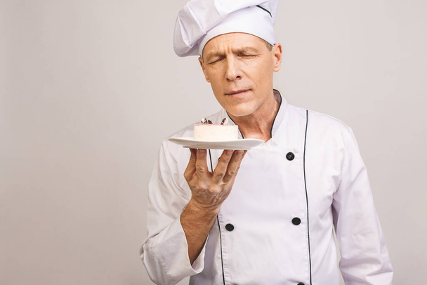 Portret szczęśliwego starszego kucharza ubranego w mundur trzymającego talerz z kawałkiem ciasta i patrzącego na kamerę odizolowaną na szarym tle. - Zdjęcie, obraz