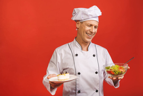 Photo de heureux jeune chef cuisinier en uniforme debout isolé sur fond de mur rouge, tenant salade et gâteau. Concept de régime alimentaire
.  - Photo, image