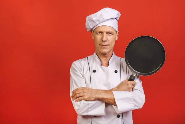 Εικόνα του χαρούμενου αρχικελευστή με στολή μάγειρα χαμογελώντας και κρατώντας το τηγάνι απομονωμένο πάνω από το φόντο του κόκκινου τοίχου.  - Φωτογραφία, εικόνα