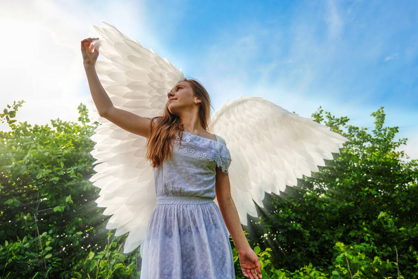 天使の羽を持つ可愛い白人女の子と青い空を背景にポーズをとった白いドレス. - 写真・画像