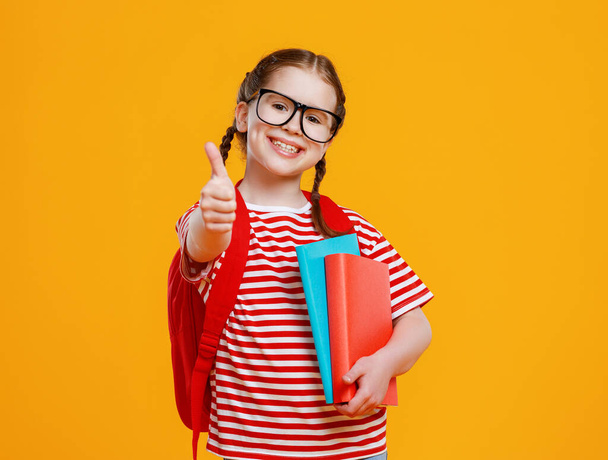 Menina inteligente positivo em óculos e com livros didáticos sorrindo para câmera e gesto polegar para cima durante os estudos da escola contra backgroun amarelo
 - Foto, Imagem