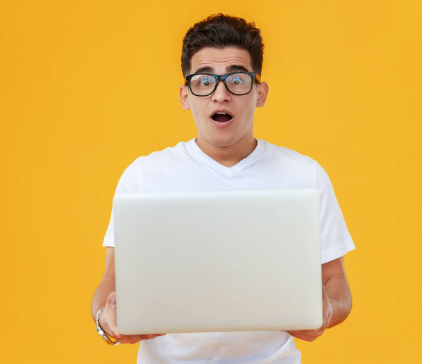 Verbaasde etnische man in wit t-shirt en bril gericht op laptop scherm met geopende mond tegen gele backgroun - Foto, afbeelding