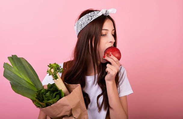 Portrait of a cute caucasian girl vegetarian woman hipster, enjoys eating fresh tasty red apple and holds green vegetables from an eco farm, lifestyle,diet in joy,healthy food. In basic white t-shirt. - Foto, Bild