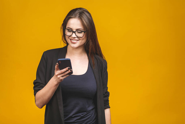 Portrait of smiling business woman in eyeglasses holding smartphone and looking back over yellow background. Using phone.  - Photo, Image