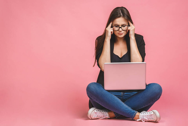 Сердита сумна нудна жінка в стресі, що сидить на підлозі з ноутбуком ізольована на рожевому фоні
. - Фото, зображення