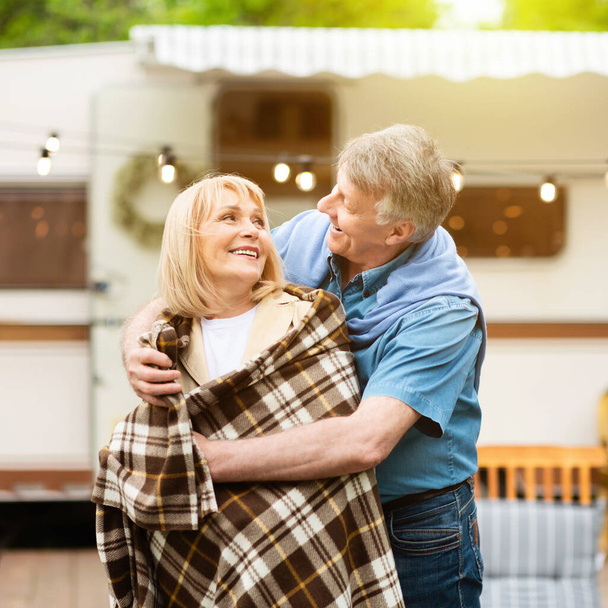 Affectionate elderly man putting checkered blanket on his wifes shoulders near trailer outdoors - Photo, Image