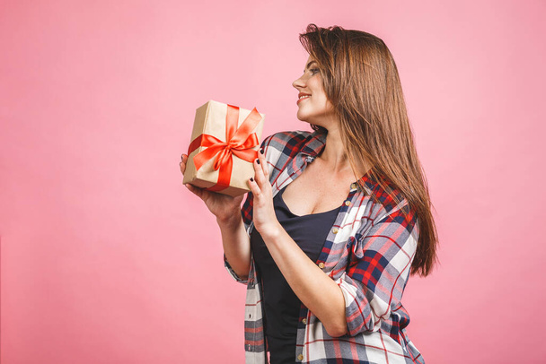 Happy young woman in casual holding gift box and looking aside while enjoys over pink background - Photo, Image