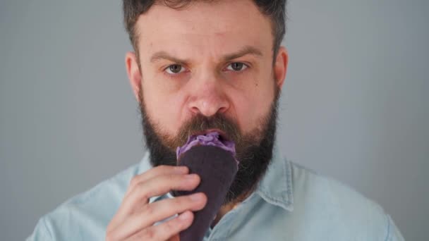 Κοντινό πλάνο ενός γενειοφόρου άντρα που τρώει μωβ παγωτό σε μαύρο κύπελλο βάφλας σε γκρι φόντο τοίχου - Πλάνα, βίντεο