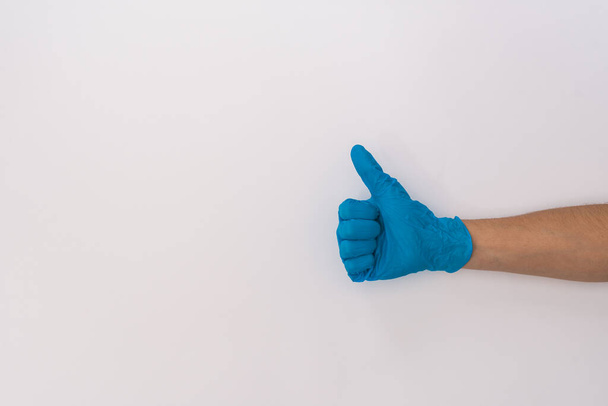 Stock photo of someones hand with blue medical latex gloves doing thumbs up on a white background. The person is unrecognizable. - Photo, Image