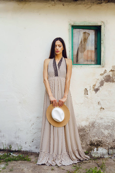One adult caucasian woman standing in front of the white wall of old traditional house wearing dress looking down sad holding hat alone loneliness solitude and grief concept - Photo, Image
