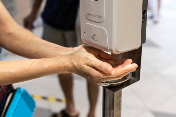 Persoon ontsmettende handen met vloeibare gel van ontsmettingsmachine in winkelcentrum - Foto, afbeelding