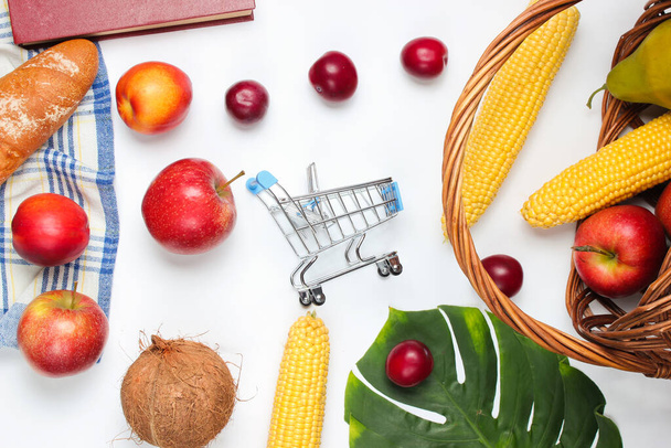 Comprar comida para un picnic. Mini carrito de compras con frutas y cesta sobre fondo blanco. Hoja de Monstera
. - Foto, imagen