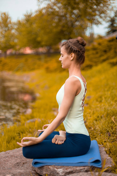 若いスポーツの女の子は、川で秋の黄色の芝生の上でヨガを練習し、ヨガのアサンスの姿勢を使用します。自然との瞑想と団結. - 写真・画像