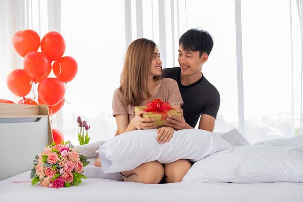 La novia se alegra de recibir una caja de regalo de oro de su novio en la cama en el dormitorio decorado con globos de corazón rojo en el Día de San Valentín
. - Foto, imagen