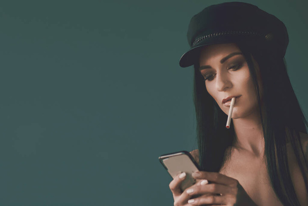 Beautiful stylish woman in tattoos, smokes cigarettes and looks into the smartphone. Connected anywhere at any time. A woman holding phone in her hands. - Photo, image