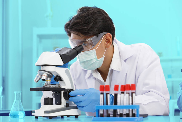 Laboratuvarda çalışan Asyalı erkek bilim adamı kan örneklerinin sonuçlarını incelerken mikroskop kullanıyor. Mavi Ton. SARS-CoV-2, Covid-19 Teması. - Fotoğraf, Görsel
