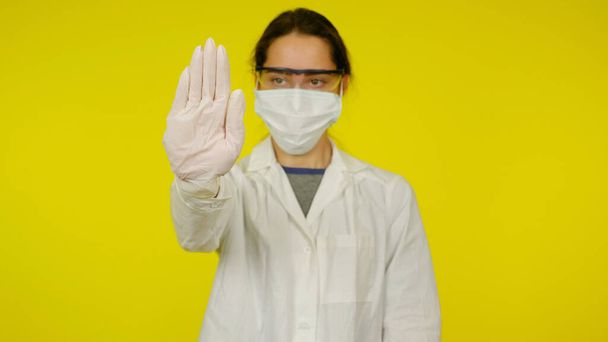 Jovem médico em uma máscara médica, óculos e luvas de látex mostra uma parada com a mão
 - Foto, Imagem