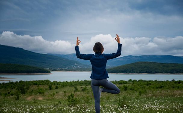 Eine junge Frau, die auf dem Feld Hatha Yoga macht, steht in Yoga-Baumpose oder Asana Vrikshasana tre Pose. Harmonie mit der Natur, Gesundheit und Fitness-Konzept. Selbstanalyse und Seelensuche - Foto, Bild