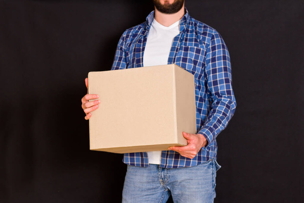 Jovem entregador de encomendas segurando uma caixa de papelão em suas mãos em um fundo preto
. - Foto, Imagem