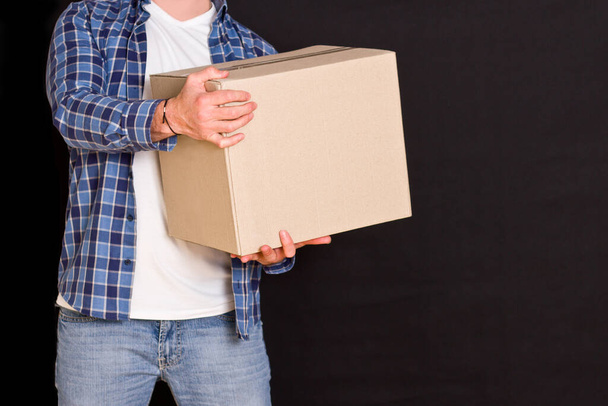 Young parcel delivery man holding a cardboard box in his hands on a black background. - Photo, Image