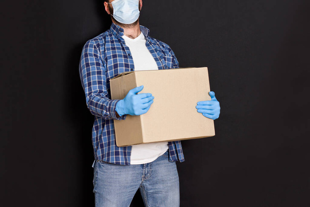 Un trabajador del servicio de entrega en una mascarilla médica que lleva una caja de cartón en la mano del mensajero con máscaras protectoras y guantes sujeta la caja de paquetes
. - Foto, imagen