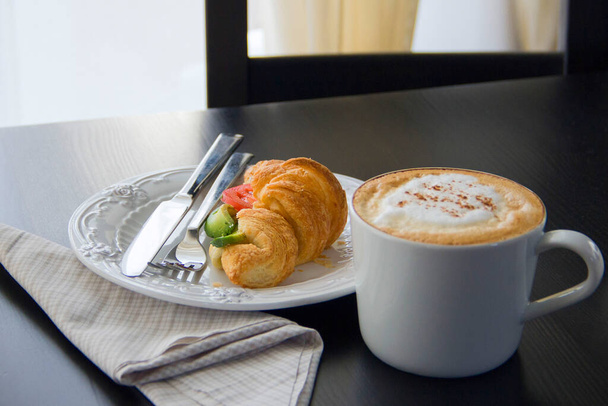 Friss croissant avokádóval és paradicsommal, egy cappuccino bögre egy sötét asztalon, reggeli fény az ablakból. - Fotó, kép