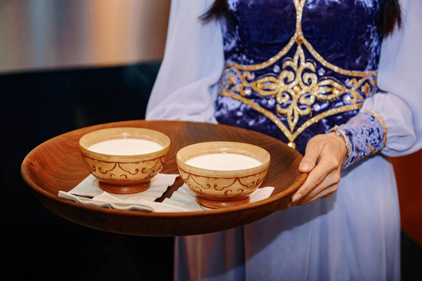 Saludo a los huéspedes con una taza de leche de caballo, tradición nacional kazaja. Primer plano de dos tazas de koumiss en un plato, con un fondo de una muestra nacional
 - Foto, imagen