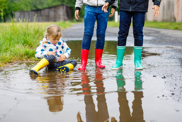 Three children, toddler girl and two kids boys wearing red, yellow and green rain boots and walking during sleet. Happy siblings jumping into puddle. Having fun outdoors, active family - Foto, Bild