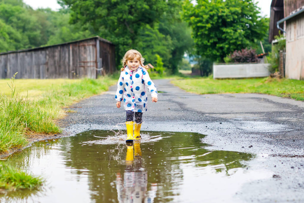 Pikkulapsi tyttö yllään keltainen sadesaappaat, käynnissä ja kävely aikana ränsistynyt sateinen pilvinen päivä. Söpö onnellinen lapsi värikkäissä vaatteissa hyppääminen lätäkköön, roiskuminen vedellä, ulkoilu - Valokuva, kuva