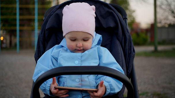 Upset little girl in blue jumpsuit with scratch on her nose sutting in pram. Child is looking at the tablet - Photo, Image