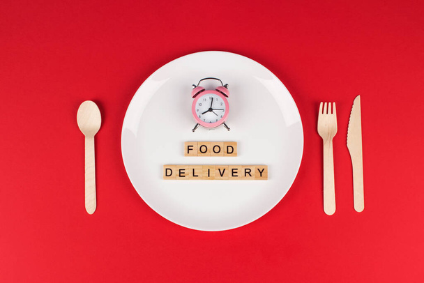 White plate, pink alarm clock, wooden cutlery set and words FOOD DELIVERY on wooden bricks on bright burgundy red background with copyspace. Creative food delivery concept, selective focus - Photo, Image