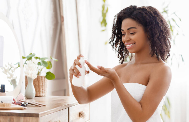 Idratante della pelle. Sorridente africano donna applicazione corpo lozione in camera da letto a casa
 - Foto, immagini