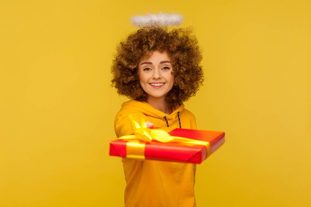 Happy holidays! Portrait of generous kind curly-haired angelic woman with saint nimbus giving wrapped present to camera, sharing christmas gifts. indoor studio shot isolated on yellow background - Photo, image