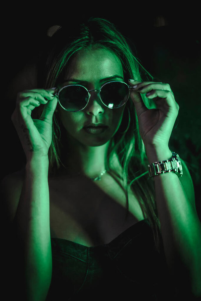 Caucasian brunette in a black dress illuminated by green led light reflected in black crystals. Urban night photography. Girl putting on sunglasses - Zdjęcie, obraz