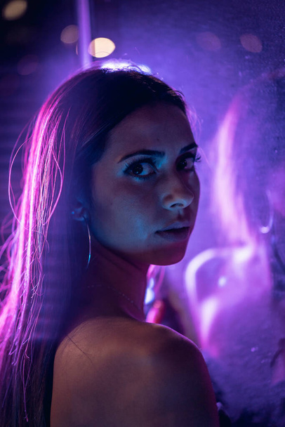 Una joven morena caucásica en un vestido negro iluminado por luz led púrpura reflejada en cristales negros, con su espalda a la cámara. Fotografía urbana nocturna
 - Foto, imagen