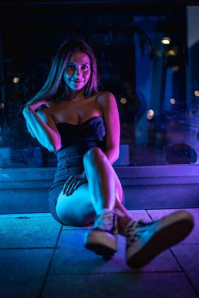 Caucasian brunette in a black dress illuminated by purple and blue led light reflected in black crystals sitting on the floor, looking to the right. Night urban photography - Foto, imagen