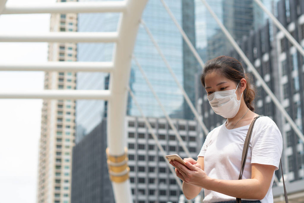 νεαρή Ασιάτισσα που φορούσε μάσκα προσώπου χρησιμοποιώντας smartphone και περπατούσε στην πόλη κατά τη διάρκεια της επιδημίας covid-19 ή coronavirus. κοινωνική απόσταση και νέα έννοια του κανονικού τρόπου ζωής - Φωτογραφία, εικόνα