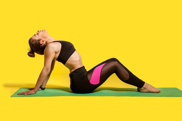 Fitness woman with hair bun in tight sportswear sitting on gym mat doing sport, bending backwards stretching spinal muscles, warming up with flexibility exercises. studio shot, isolated on yellow - 写真・画像