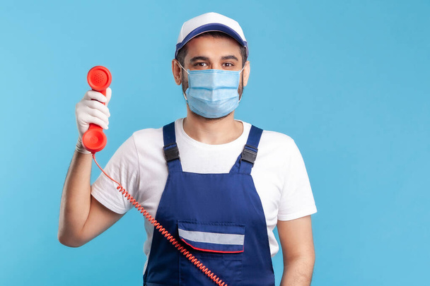 Housing maintenance call centre. Friendly handyman in overalls, mask, safety gloves, holding retro phone handset, answering customer call. Plumber and repair services. indoor studio shot, isolated - Photo, Image