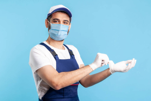 handyman in overalls and mask showing hand with surgical gloves, smiling to camera. Safety clothes, protection and hygiene in service industry, courier delivery, housekeeping maintenance. indoor. - Fotó, kép
