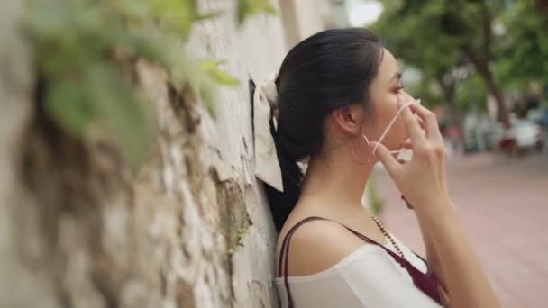 Vonzó ázsiai nő visel fehér védő maszk, oldalnézet vissza a falra, covid-19 koronavírus, környezetvédelem, nappali, új normális, szabadtéri jelenet, ázsiai hölgy fel maszk - Felvétel, videó