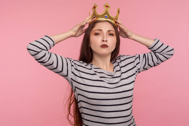 I'm leader! Portrait of ambitious successful woman adjusting golden crown on head and looking with arrogance, declaring her authority, superiority. indoor studio shot isolated on pink background - Φωτογραφία, εικόνα