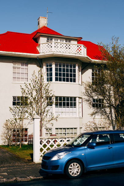 Maison de trois étages avec de grandes fenêtres panoramiques et un grenier au toit rouge à Reykjavik, la capitale de l'Islande. Une voiture bleue est garée près de la maison le long de la route
. - Photo, image
