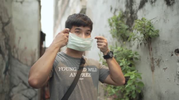SLOW motion, Asian young man wearing surgical mask, covid-19 corona virus, healthcare, Day time, new normal, outdoor scene, model looking at camera, asian male put on mask, green leaf environment  - Footage, Video