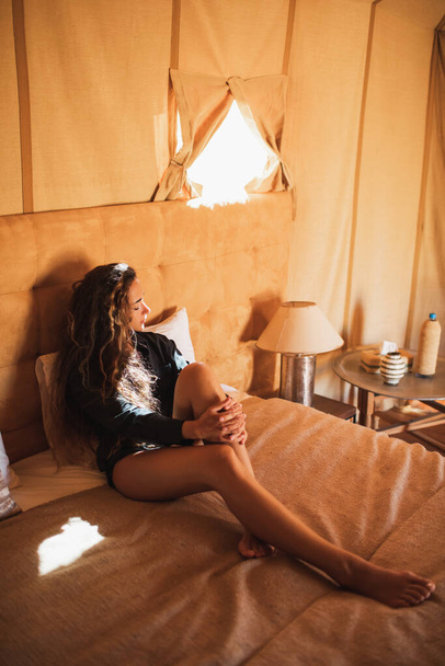 Sensuele glamour portret van oosterse vrouw in het interieur van moderne luxe glamping tent kamp in Marokko. Ontspannen op bed. Ecodesign concept, natuurlijke en houten elementen in stijl. - Foto, afbeelding