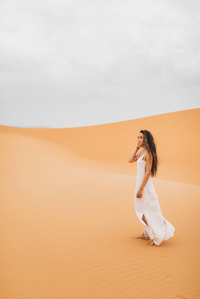 Ładny portret młodej pięknej kobiety na wydmach pustyni marokańskiej Sahary. Brunetka z długimi włosami, wschodni wygląd. Panna młoda w białej jedwabnej sukni. - Zdjęcie, obraz