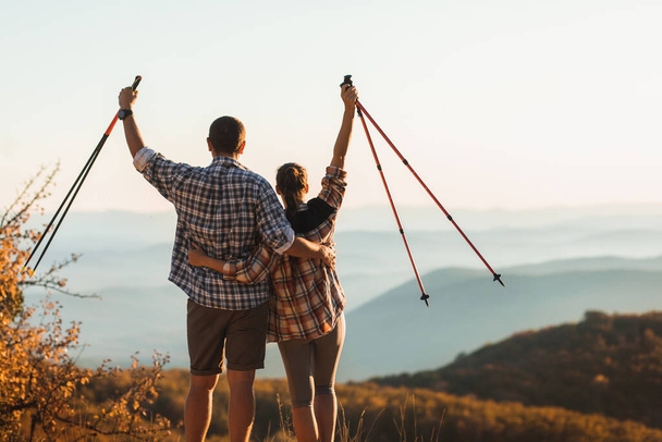 Couple happy to climb top of mountain and hold trekking sticks. Nordic walking outdoors, view from behind. Travel lifestyle. Success goal achievement. - Photo, Image