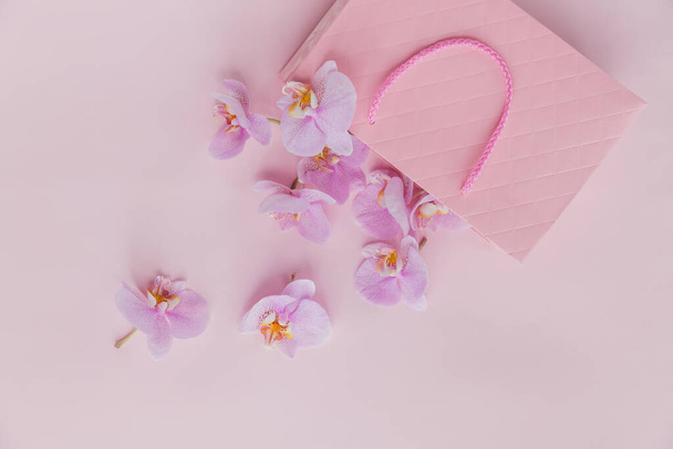 Pink gift bag and flying orchid flowers on light pink background. Top view greeting card with delicate flowers. Holiday, Women's day, mother's day greeting concept. - Photo, image