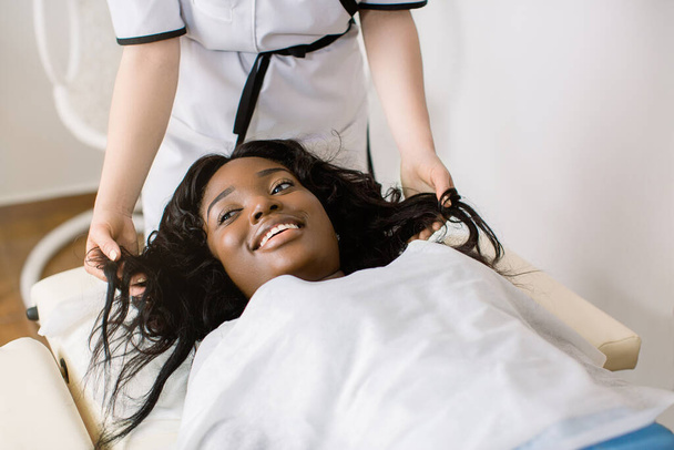 Cropped image of hands of female doctor cosmetologist trichologist dermatologist examining the state of the patients hair and looking at damaged hair tips. Smiling African girl lying on the couch - Zdjęcie, obraz