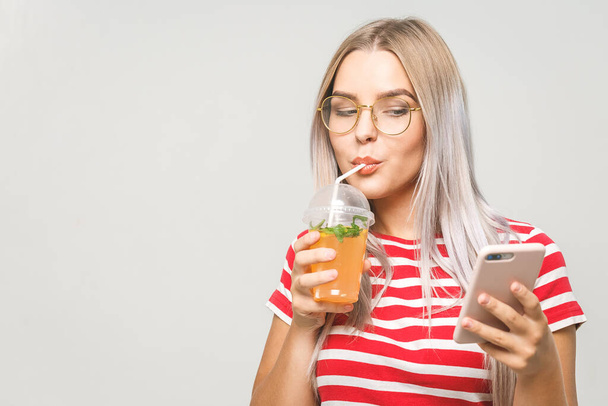 Beautiful young woman drinking a homemade detox juice, texting on her phone while isolated over white. Using phone.  - Фото, изображение